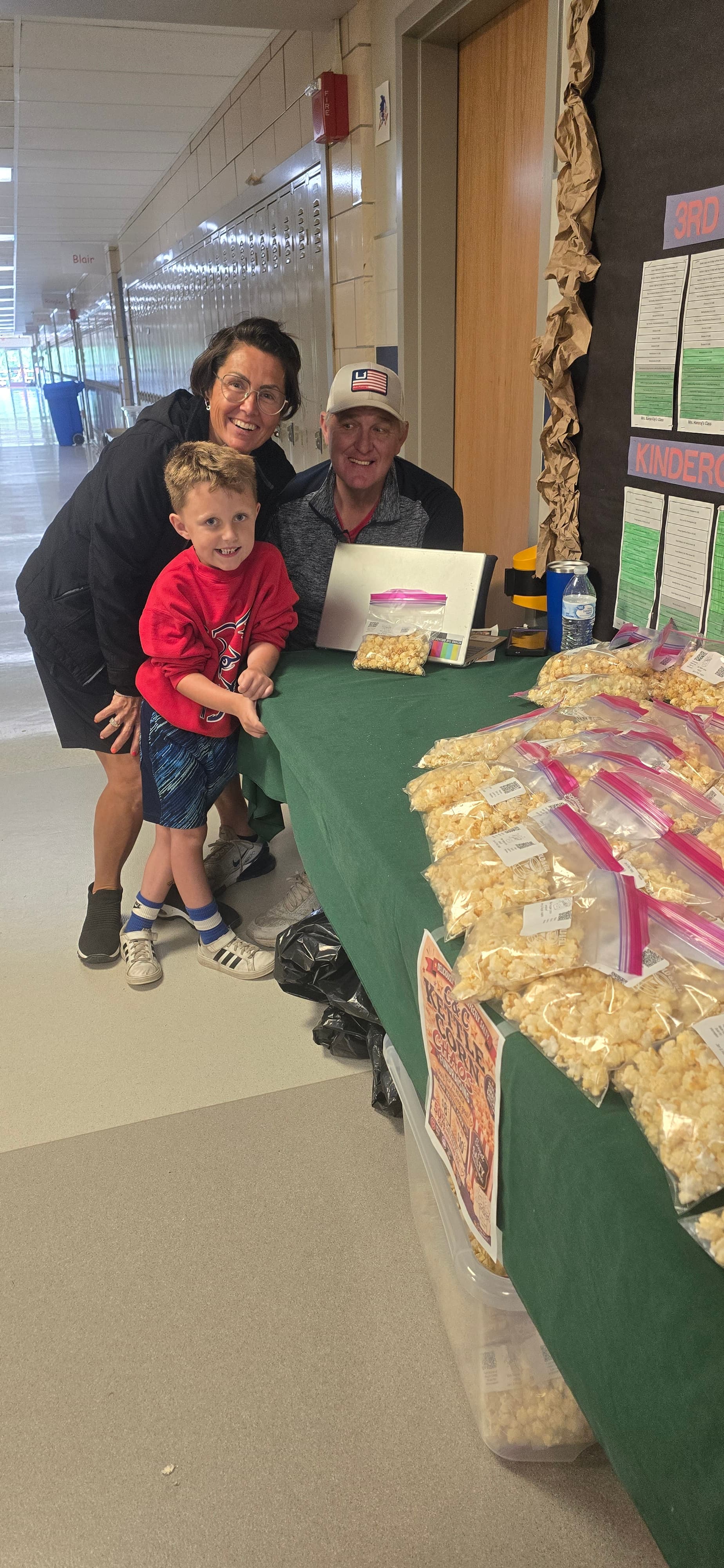 Woman, man, and boy smile behind a table filled with bags of popcorn.